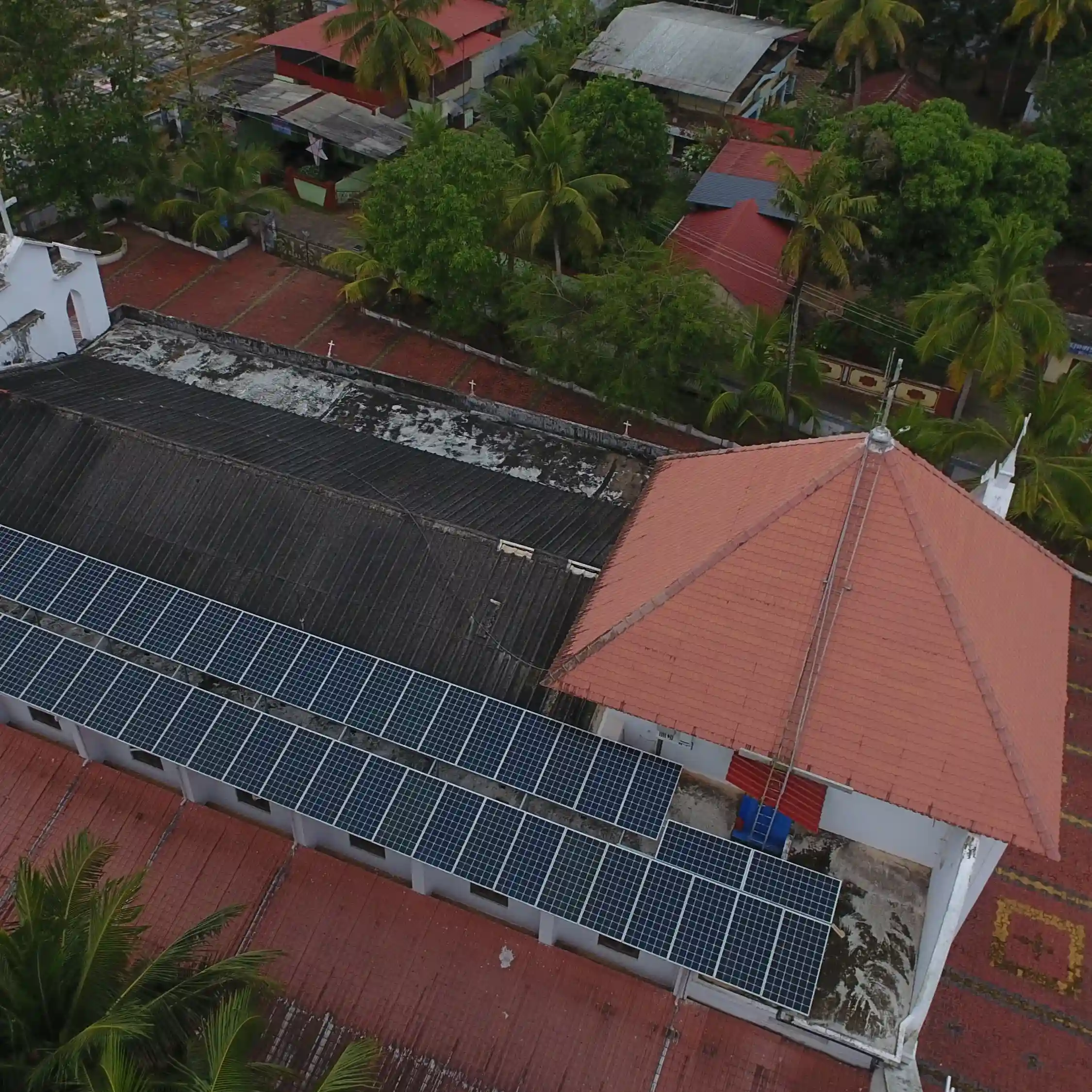 Aerial view of a white church with red roofs and a bell tower, featuring solar panels on the roof. Surrounded by trees and colorful umbrellas in a courtyard.