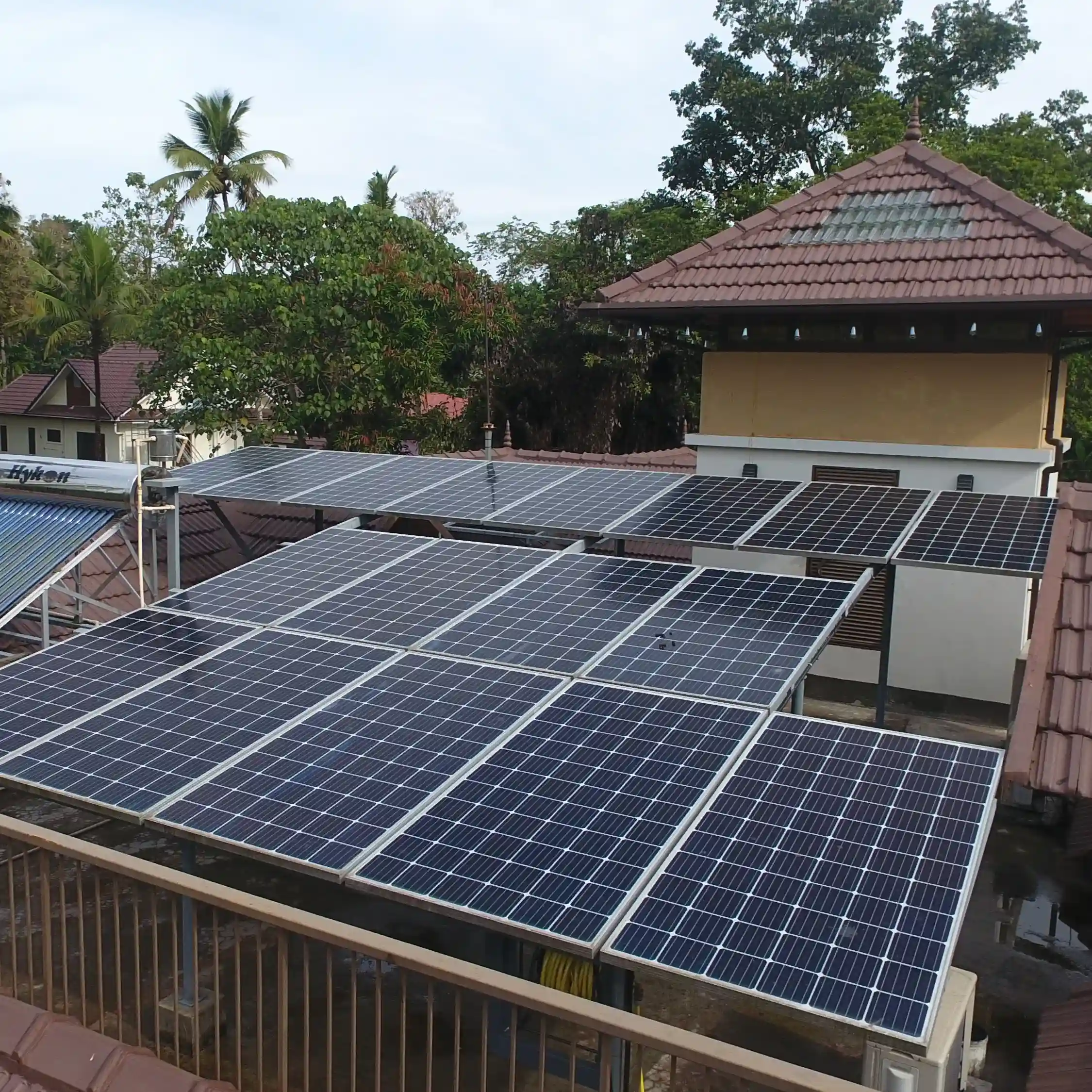A rooftop covered with solar panels and a solar water heater, surrounded by trees and tile-roofed structures, conveying a sense of sustainable living.