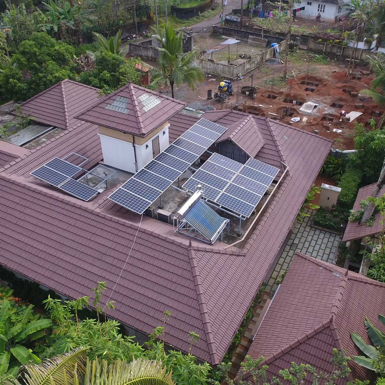 Aerial view of a house with solar panels on a brown tiled roof, surrounded by lush greenery. A construction site is visible in the background.